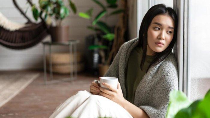 Sad asian woman looking outside window sitting on floor with miserable upset face drinking coffee at...