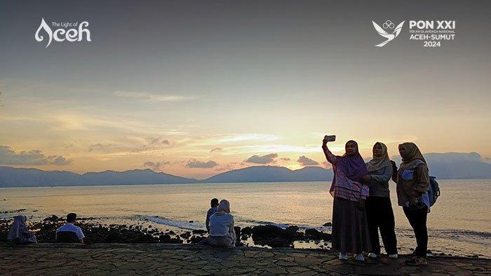 Pengunjung mengabadikan momen dengan berfoto selfie di tepi Pantai Sabang Fair saat matahari terbenam, dengan latar pemandangan laut yang indah dan gunung yang tampak samar di kejauhan.
