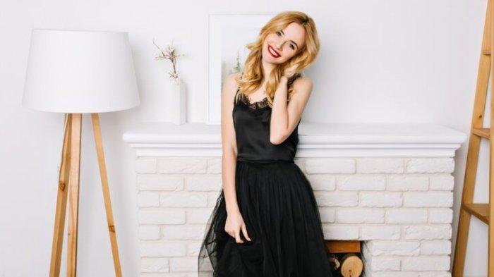 Beautiful young lady wearing elegant black dress in white room, posing, smiling. She's touching her long beautiful wavy golden hair.
