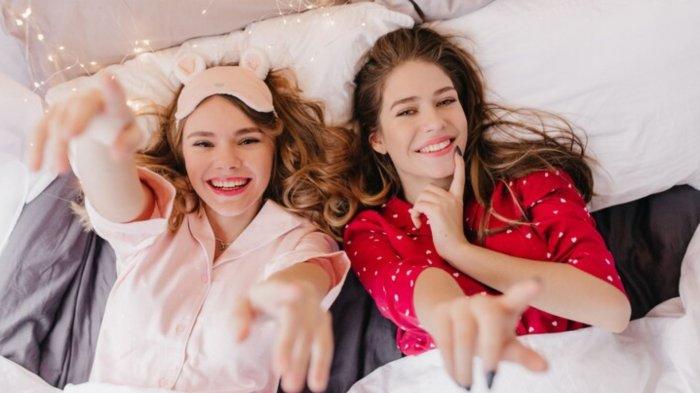 Overhead portrait of two winsome girls lying in bed and smiling. Curly woman in pink eyemask posing with pleasure in morning.