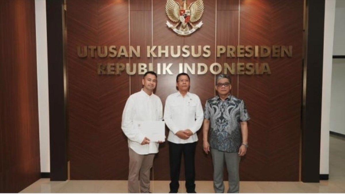 Raffi Ahmad yang mengenakan kemeja putih dan celana panjang cokelat berpose di depan dinding berhiaskan lambang Garuda dan tulisan 