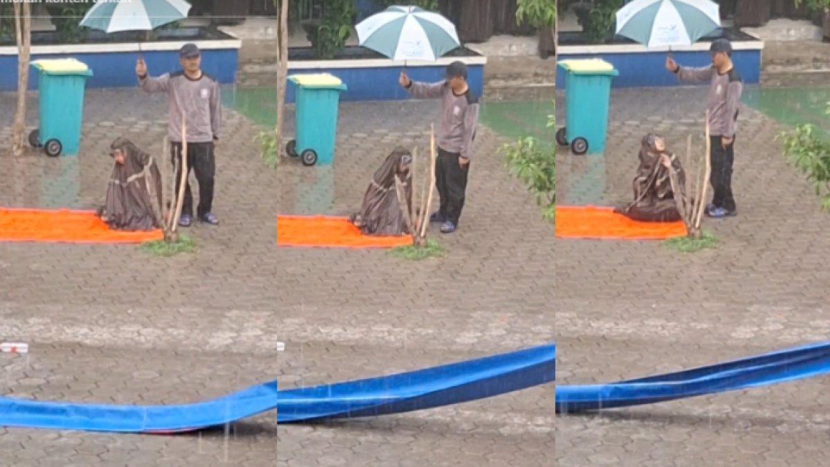 Seorang satpam rela basah kuyup demi memayungi guru yang sedang salat ketika hujan mendadak datang