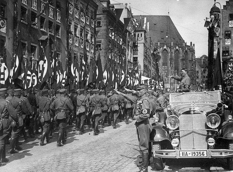 Adolf Hitler berdiri di dalam Mercedes-Benz 770- nya meninjau anggota SA ( Sturmabteilung ) dalam parade selama rapat umum Nuremberg 1935, Kongres Partai Nazi ke-7yang diadakan di Nuremberg, 10–16 September. Hitler didampingi oleh Blutfahne , ('Bendera Darah', bendera swastika seremonial) dan pembawanya SS-Sturmbannführer Jakob Grimminger. File gambar ditandai sebagai domain Publik di Narodowe Archiwum Cyfrowe , Arsip Digital Nasional Polandia.
