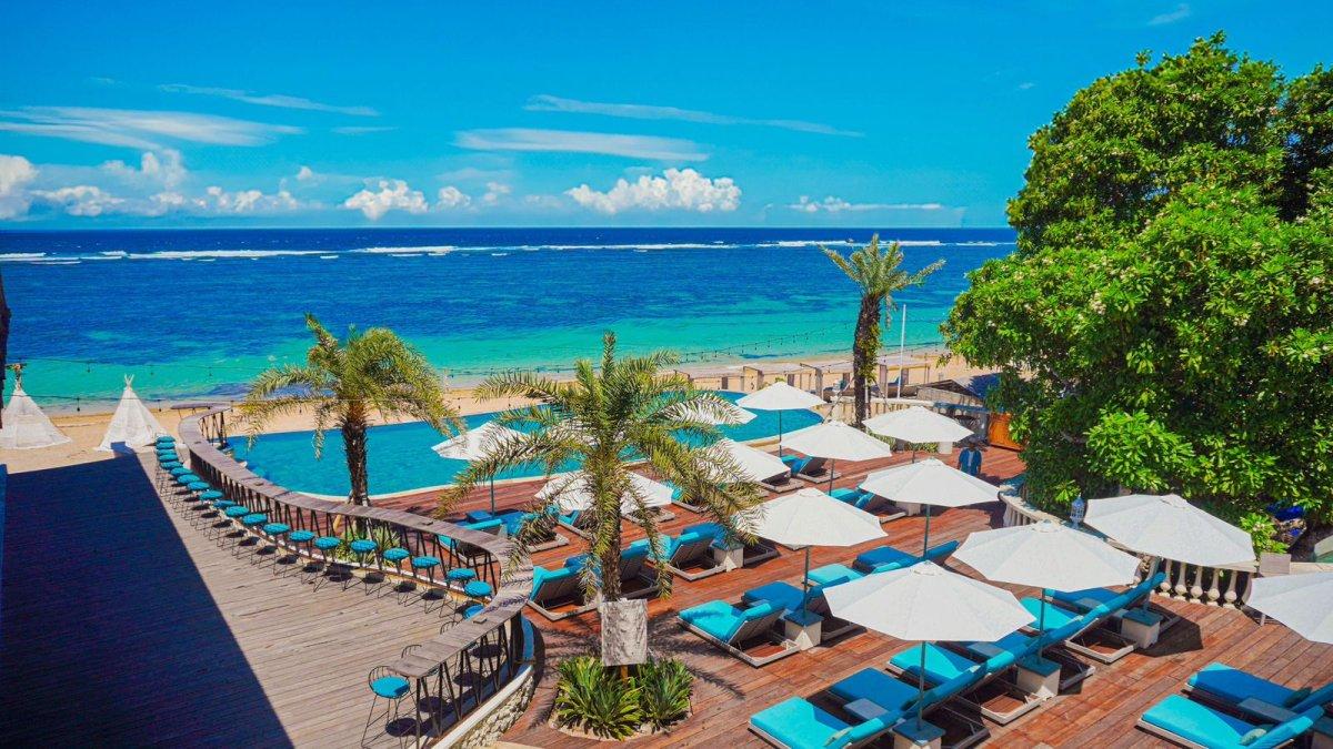 Kolam renang dan area santai tepi pantai di CANNA Bali, Nusa Dua — tempat ideal untuk menikmati suasana laut biru, dengan fasilitas sunbed dan bar outdoor yang menyatu dengan pemandangan tropis.