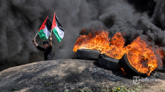 Seorang pria memegang bendera Palestina saat dia berdiri di dekat asap dari ban yang terbakar selama demonstrasi di sepanjang perbatasan dengan Israel di sebelah timur Kota Gaza pada 5 April 2023. - Polisi Israel mengatakan mereka telah masuk untuk mengusir